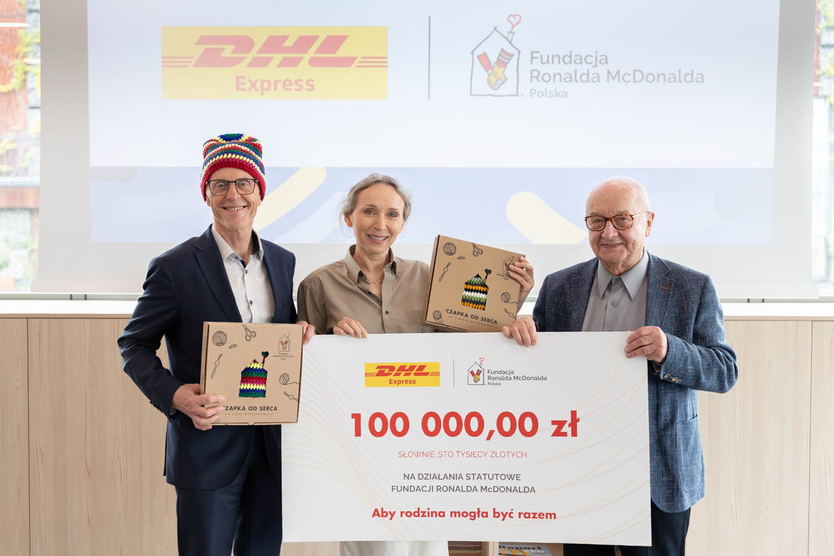 Symboliczny czek na 100 tys. zł od DHL dla Fundacji Ronalda McDonalda – na zdjęciu trzy osoby zaangażowane w projekt „Czapka od serca”.